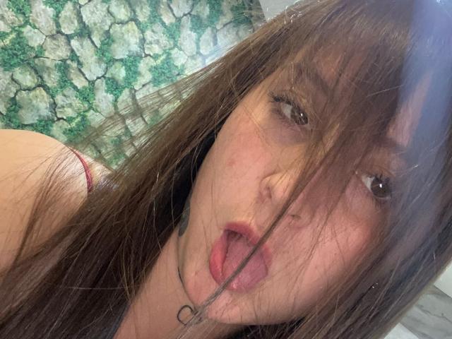 EvaDaMore with brown hair, slightly open mouth, tongue out, and red bra strap visible, wearing a black choker, brown eyes, and messy hair in a selfie.