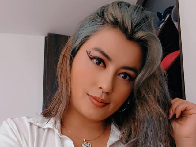 BellaBlue69 has long, wavy, greyish-brown hair, thick black eyeliner, a nose ring, and is wearing a white shirt, looking at the camera with a slight smile.