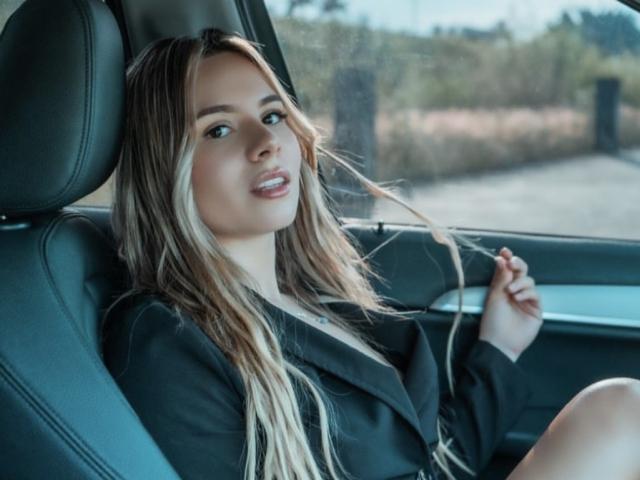 AnaMazzeo with long blonde hair, black blazer, and light skin, sitting in a car, holding her hair, smiling, showing her teeth, with a relaxed, sexy vibe.
