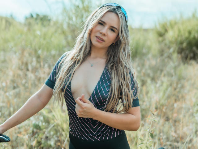 AnaMazzeo with long blonde wavy hair, wearing a deep V-neck striped top, exposing her small tits, smiling, holding a bike, blue sunglasses on head, outdoors, sunny day.