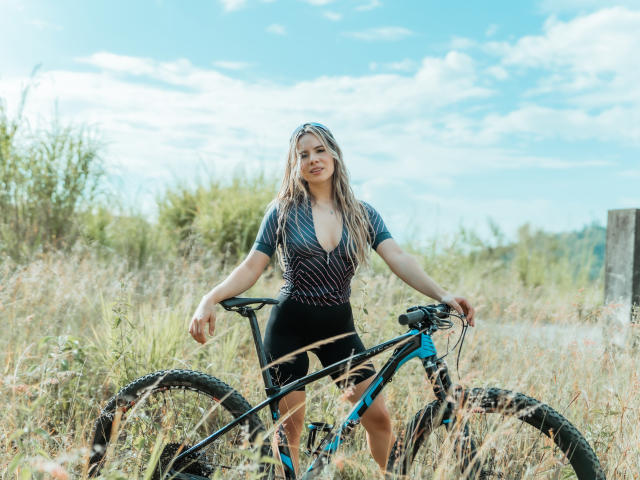 AnaMazzeo with long blonde hair, wearing a deep V-neck, black and gray bike outfit, standing in grassy field, holding blue and black mountain bike, smiling.