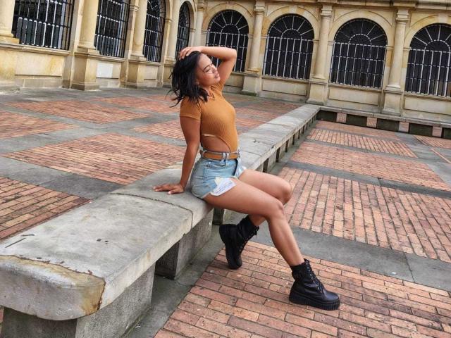 SoleneFontaine sits on a concrete bench, wearing a tan crop top, blue denim shorts, white underwear, black boots, and a belt. She's posing with her hand in her hair.