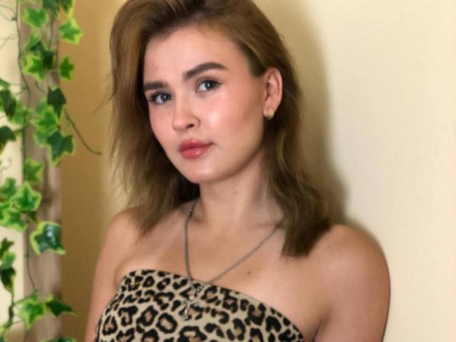 SofiVernara with shoulder-length brown hair, wearing a leopard-print strapless top, has pink lips, blue eyes, and a silver necklace, standing against a yellow wall with green vines.