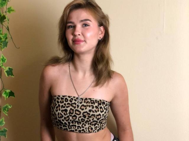 SofiVernara with light brown hair, wearing a leopard print strapless top, showing her small boobs, a silver necklace, and a slight smile, standing against a yellow wall.