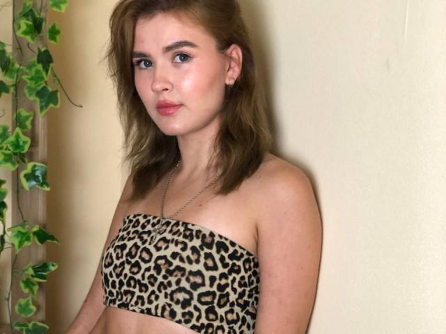 SofiVernara mit hellbraunen, gewellten Haaren, trägt ein trägerloses Top mit Leopardenmuster, das kleine Brüste zeigt, und steht vor einer beige farbenen Wand, wobei sich links grüne, blättrige Ranken befinden..