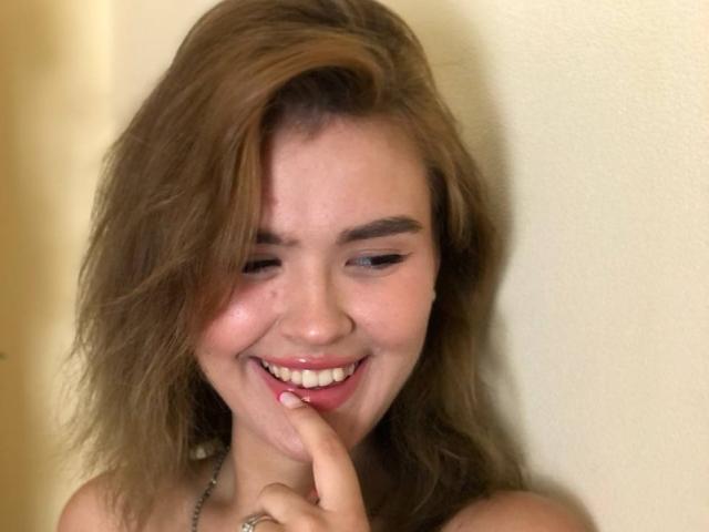 SofiVernara with wavy brown hair, smiling, holding finger to lips, showing white teeth, and wearing a necklace, has a cute face and is looking down.