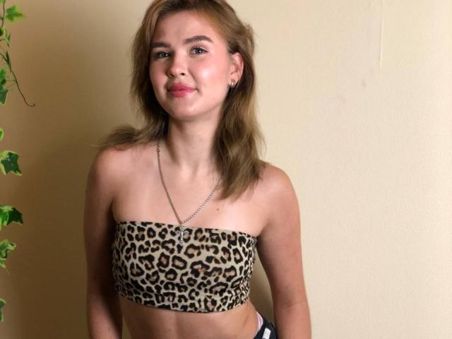 SofiVernara with light brown hair, wearing a leopard print strapless top, showing her small tits, smiling, and wearing a chain necklace, against a plain yellow wall.
