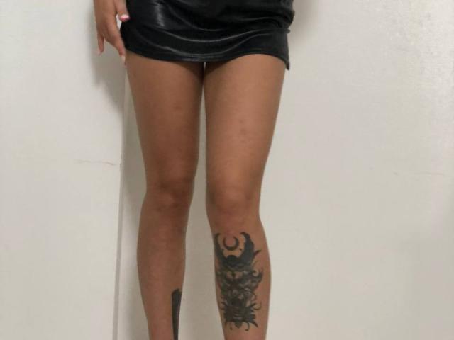 Las piernas de SofiVernara, vestida con una minifalda de cuero negro, mostrando un tatuaje de un diseño oscuro y elaborado en su muslo derecho, de pie frente a una pared blanca..