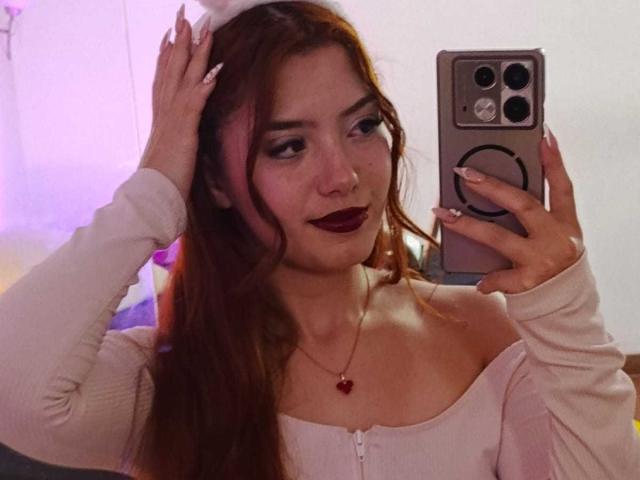 EmmaThomsonn with long brown hair, wearing a white off-shoulder top, dark lipstick, and a red necklace, taking a selfie with her phone, hand in hair, smiling.