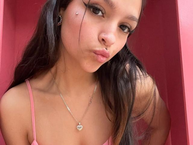 MarietteJones has long dark hair, a nose ring, and a heart necklace, pouting with full lips, wearing a pink top, showing her small tits, and looking seductive.