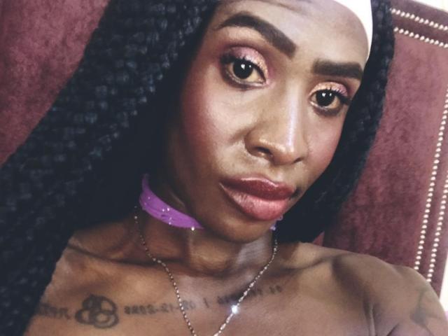 SamaraaJackson69 with dark skin, long black braids, pink choker, shiny red lips, brown eyes, small nose, tattoos on chest, necklace, and serious expression.