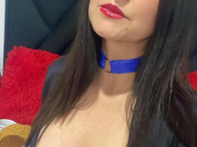 KynkyFetish tiene labios rojos brillantes, cabello negro lacio, y lleva un collar azul con un anillo, mostrando su escote y su piel clara en un primer plano..