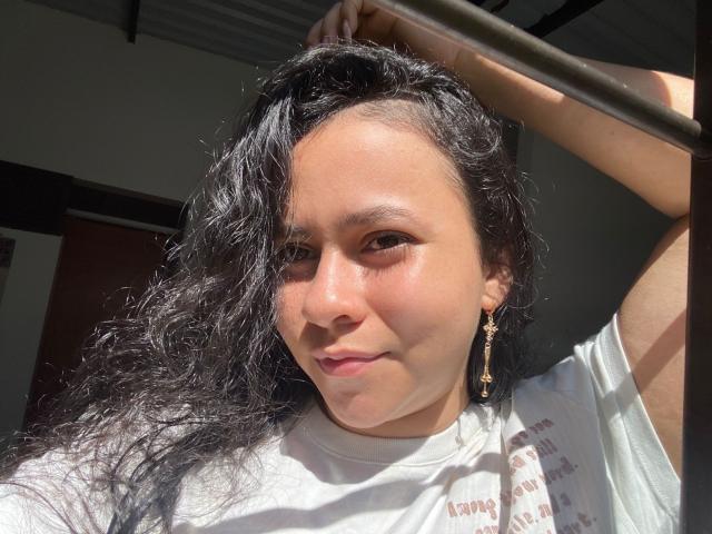 SamanthaLorr with curly black hair, sunlight on her face, white shirt, gold earrings, right arm raised, relaxed smile, soft skin, slight blush, casual, confident, natural light, close-up.