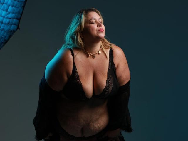 AllegraFeragamo has big tits, a soft belly, and light skin, wearing black lace lingerie with a black shawl, eyes closed, necklace on, and a blue light on the left.