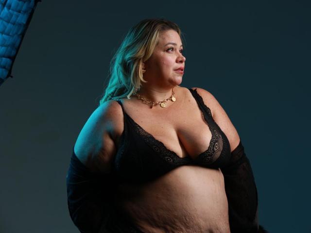 AllegraFeragamo with blonde hair, wearing black lace bra, showing her large tits and a soft belly, gold necklace, looking to the side, in a dimly lit studio.