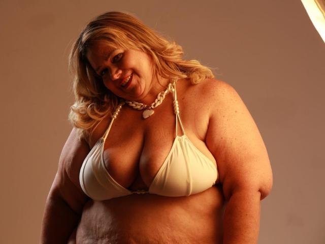 AllegraFeragamo, plus-sized woman with blonde hair, wearing a white bikini top, showing large boobs, smiling, with a white necklace, standing in warm light, looking down.