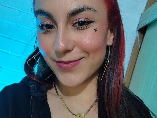 AmyStreetwise69 has red-tipped black hair, eyeliner, and a small black star tattoo near her right eye. She's smiling with pink lipstick, wearing gold earrings and a black jacket.