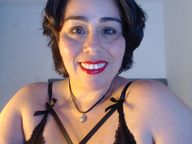 AnastaciaBlues met kort bruin haar, rode lippenstift, een zwarte kanten bh en een ketting, lacht naar de camera en laat haar borsten zien. Ze is een schattige, mollige vrouw met een sexy glimlach..