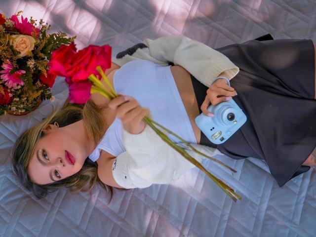 EmilyVelours lying on a quilted bed, holding yellow flowers, wearing a white top, black skirt, and white cardigan, with a blue camera in her other hand. Bouquet nearby.