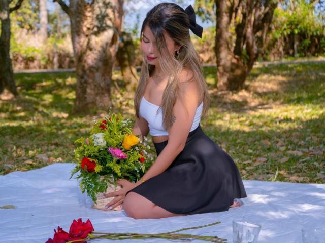 EmilyVelours has long brown hair, a white strapless top, and a black skirt, kneeling on a white blanket, holding a colorful bouquet, with red roses in front.