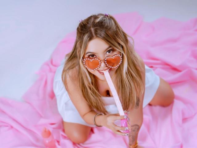 EmilyVelours on knees, white dress, pink heart-shaped glasses, sucking pink straw, blonde hair, pink fabric, tattoo on right arm, looking up, cute, sexy, pink heart-shaped straw holder.
