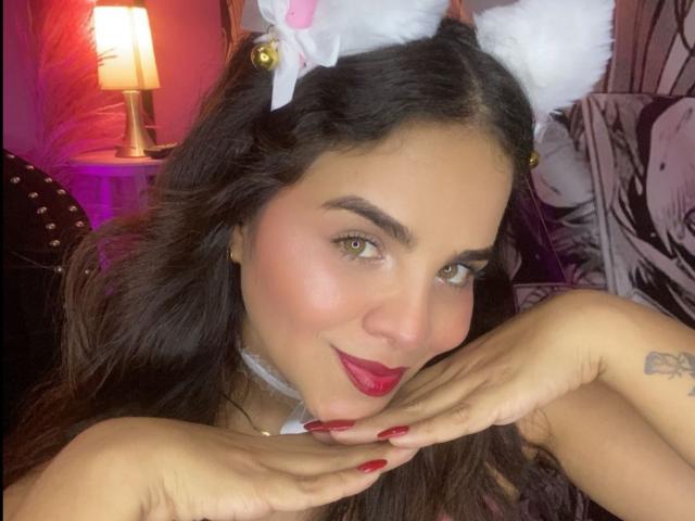 SexyNikole with cat ears, red lipstick, and manicured nails, hands near face, smiling, green eyes, white choker, tattoo on right hand, black hair, soft pink light.