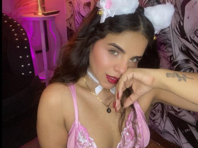 SexyNikole with dark hair, pink cat ears, red lipstick, and a pink lace top, touching her cheek, showing a tattoo on her right hand, with a seductive smile.