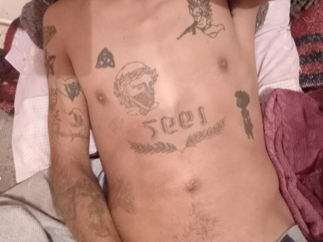 CarlitoPierre's tattooed, shirtless torso with chest and arm tattoos, including a number, leaf, and silhouette, lying on a bed with red and white pillows.