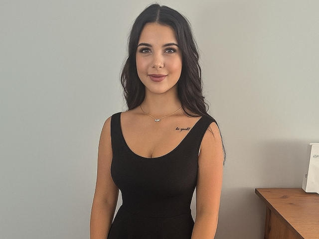 TaliaAttractive with long dark hair, wearing a black tank top, showing her tits, small tattoo on right side of chest, standing against a plain wall, smiling slightly.