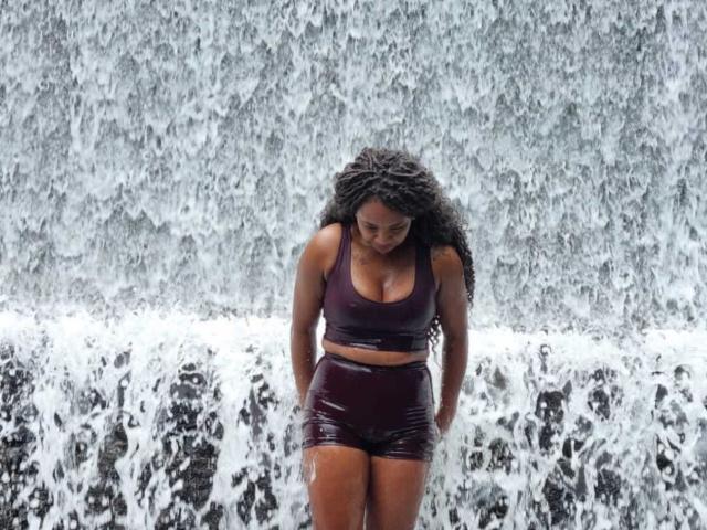 AngelaTammy with curly black hair, wet burgundy sports bra, and matching shorts, standing under a waterfall, looking down with a slight smile, water cascading around her.