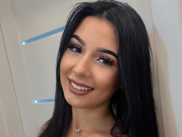 TaliaAttractive has long black hair, dark eyebrows, and is smiling with a nose piercing and lip piercing. She's wearing a silver necklace with a small butterfly pendant.