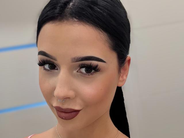 TaliaAttractive has black hair, a nose piercing, a septum piercing, and a lip piercing. She's wearing dark lipstick and has thick, long eyelashes. She's looking at the camera.