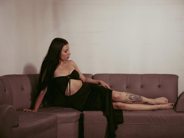 LizzeJoy has long black hair, wearing a black dress with straps, showing her tits and a thigh tattoo, lying on a purple couch, legs out, looking down.
