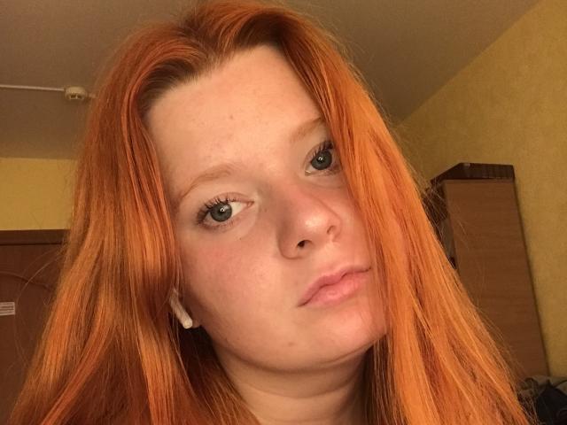 PollyKatie has long red hair, blue eyes, and a nose piercing. Her face is slightly tilted with a neutral expression. She's wearing white earbuds.