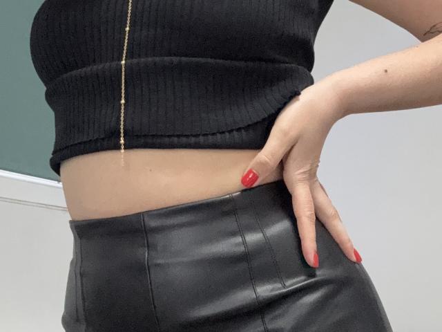 EricaLust's black ribbed crop top, showing her boobs, with black leather pants and red nails, hand on hip, gold chain, and a tattoo on her right arm.