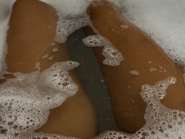 EricaLust's legs in a bathtub, brown skin, soap bubbles covering her thighs, and a metallic bath tool between her legs. Her legs are partially submerged in water.