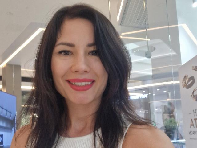 Markisa8 with dark brown, wavy hair, smiling with red lipstick, white sleeveless top, and visible breasts, in a brightly lit indoor setting.
