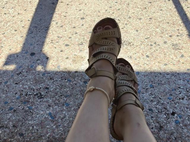 Las piernas de Dreia, con sandalias marrones de punta abierta, con pequeños agujeros y un brazalete en el tobillo, están sobre un pavimento áspero y bañado por el sol, proyectando una sombra. Lleva un tobillero beige..