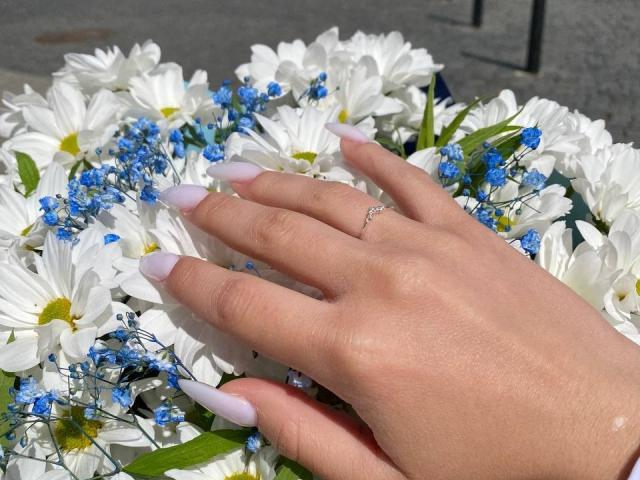 Kripkoa's hand with long, pale pink nails rests on a bouquet of white daisies and tiny blue flowers, showing a silver ring on the ring finger.