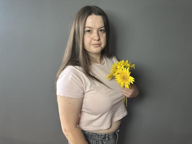 Mavka, avec de longs cheveux bruns, un t-shirt rose clair et un jean bleu, tenant un tournesol jaune, debout devant un mur gris, montrant sa petite poitrine et un sourire discret..