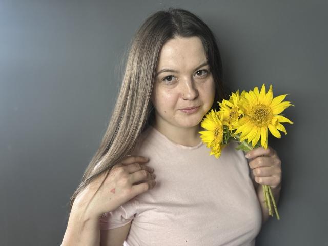 MavkaCute with long brown hair, pink shirt, holding bright yellow sunflowers, slight smile, gray background, right hand touching shirt, left hand holding flowers.