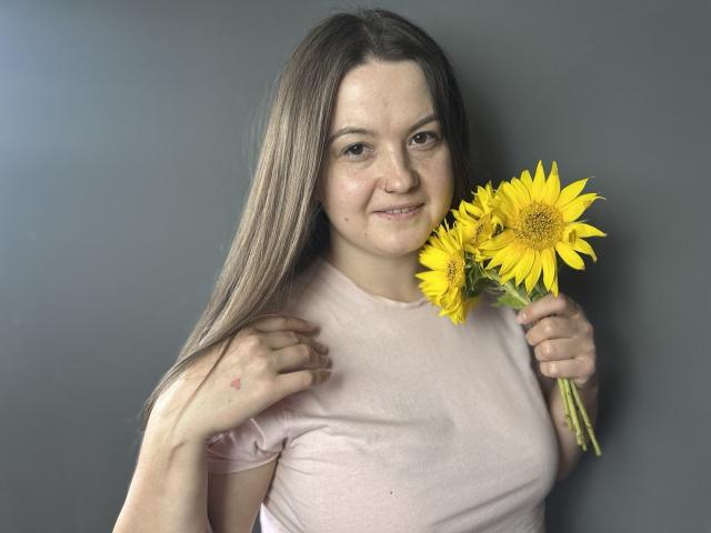 MavkaCute with long brown hair, pink shirt, holding sunflowers, slight smile, gray background, right hand touching shirt, left hand holding flowers, slight freckles, natural light, casual.