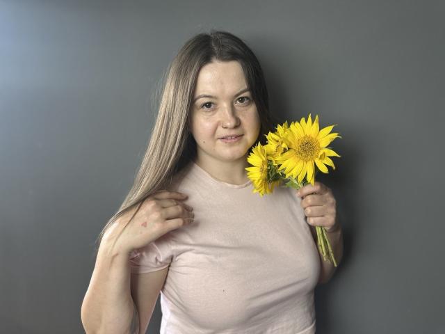 MavkaCute with long brown hair, light pink shirt, holding yellow sunflowers, slight smile, gray background, right hand touching hair, left hand holding flowers.