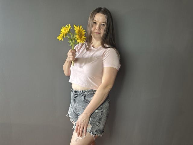 MavkaCute with yellow sunflowers, wearing a pink crop top and frayed denim shorts, standing against a gray wall, holding flowers in her right hand.