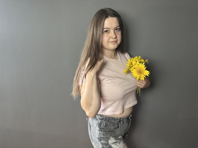 Mavka: Uma jovem com longos cabelos castanhos, uma blusa cropped rosa e calças jeans cinzas, segurando um girassol amarelo, em pé em frente a uma parede cinza, sorrindo levemente, mostrando seus pequenos seios..