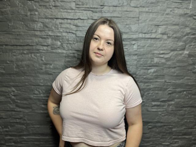 MavkaCute with long brown hair, light pink shirt, small tattoos on arms, standing against textured gray wall, slight smile, visible boobs, soft light, casual pose.