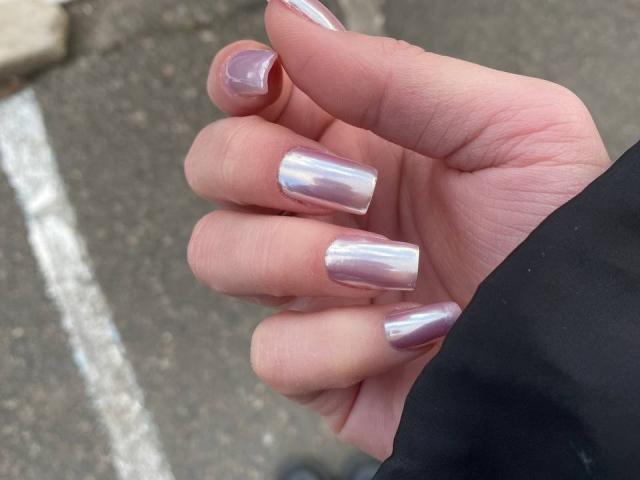 Kripkoa's hand with shiny, metallic pink nail polish on medium-length nails, showing a reflective, almost holographic finish. The skin is pale with visible knuckles.