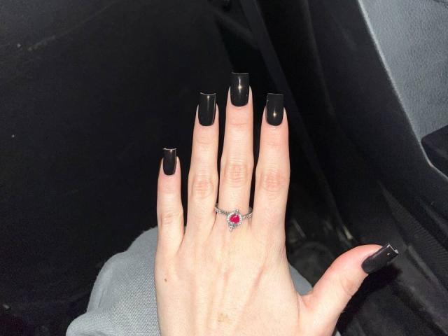Kripkoa's hand, met zwarte, extra lange, vierkante nagels, draagt een rode edelsteenring op de middelvinger, in een donkere auto. De borsten zijn niet zichtbaar..