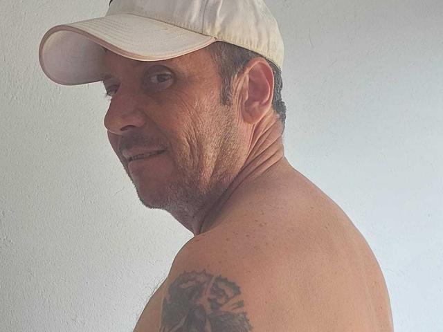 Michaell69, shirtless with a white cap, showing a tattoo on his right shoulder. His face is slightly wrinkled, and he's looking over his shoulder with a subtle smile.