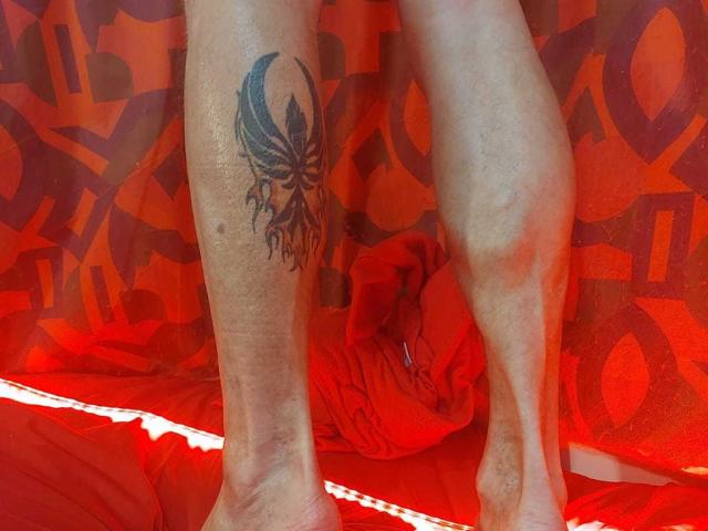 Michaell69's leg with a black, tribal-style tattoo of a butterfly on his upper thigh, standing on a red, patterned surface with Michaell69's bare, hairy feet visible.